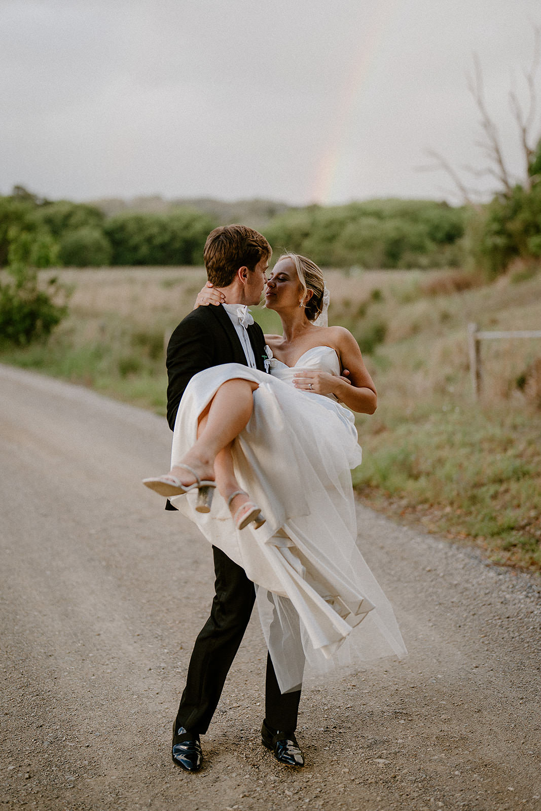 A photo of a handsome groom picking up his bride on a country road with a rainbow in the background at their byron bay wedding.