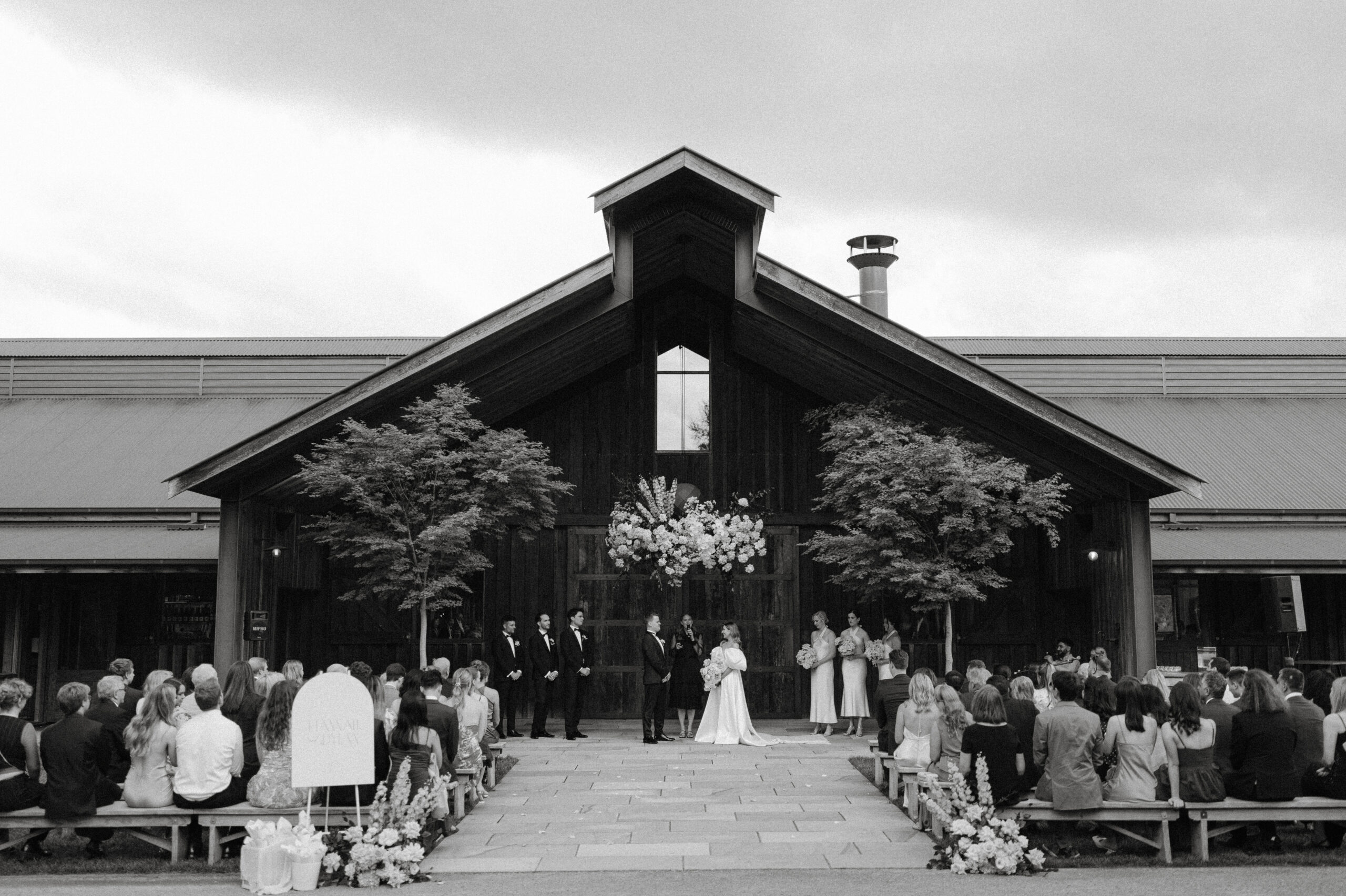 A wide photo showing a ceremony taking place under the bendooley stables architectural A Frame.